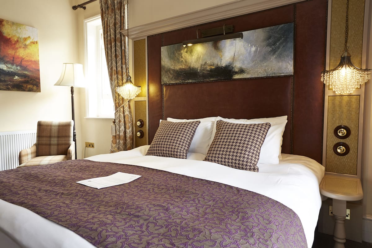 Habitación de hotel con una cama grande, cabecera con pintura, mesita de noche con lámpara y vistas al exterior.
