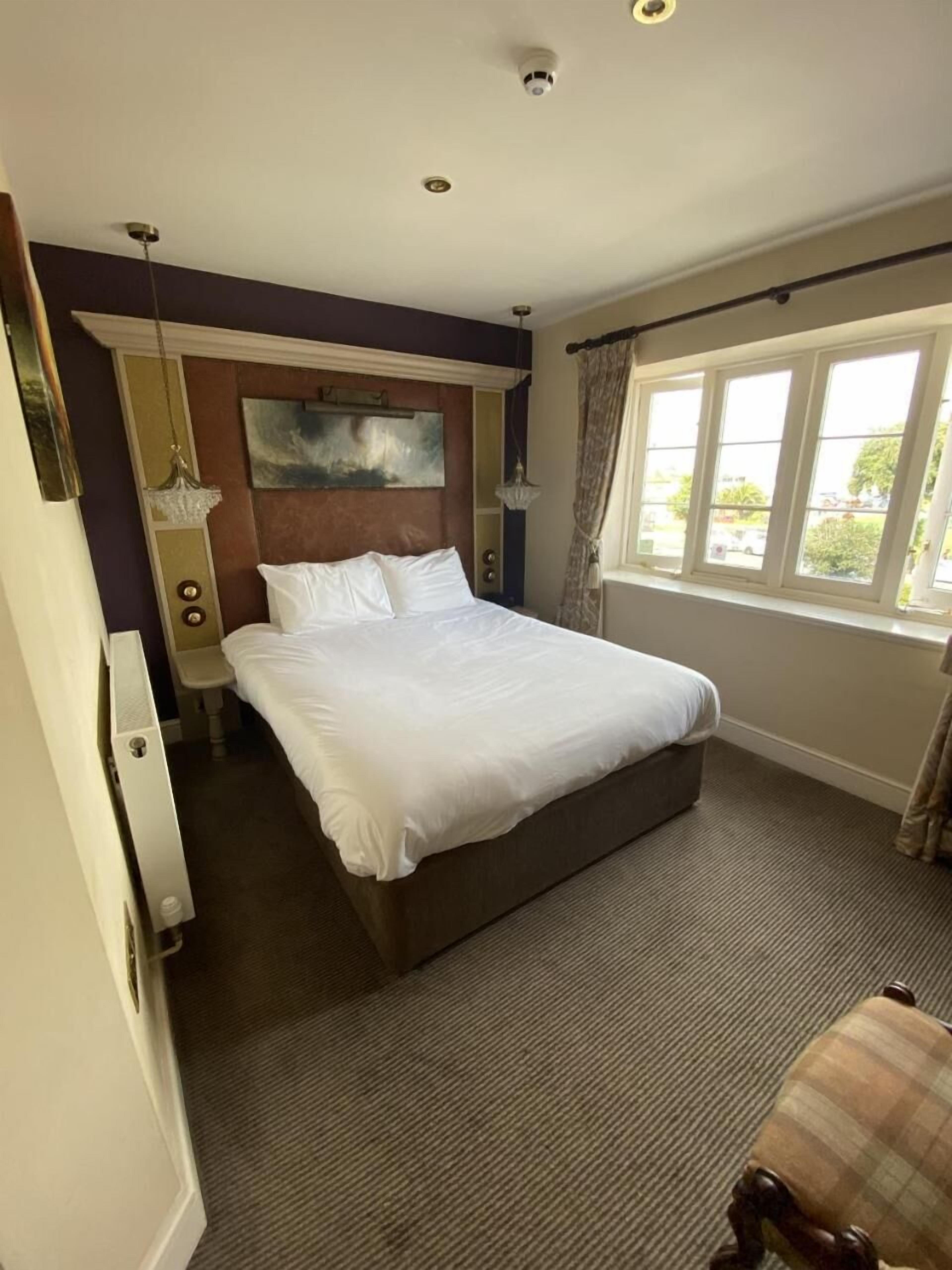 Una camera d'albergo con un grande letto, un comodino, una sedia, una finestra con tende e un quadro appeso al muro.