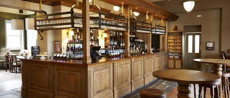 A bar area with wooden bar counter, shelves with bottles, glasses, and a menu sign. There are stools and tables around the bar.