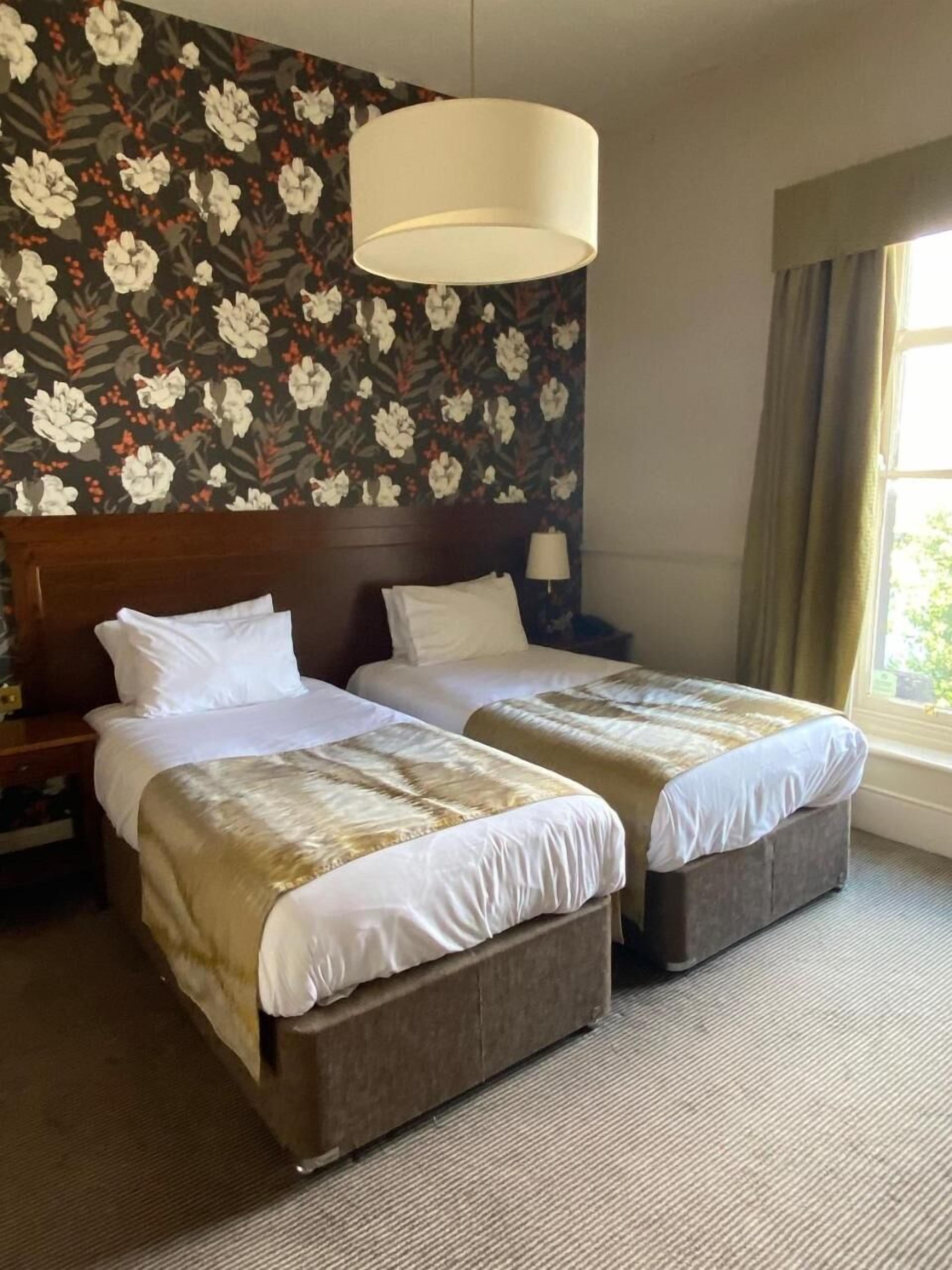 Una habitació d'hotel amb dos llits, una paret amb un gran disseny floral, una làmpada penjant i una finestra amb cortines.