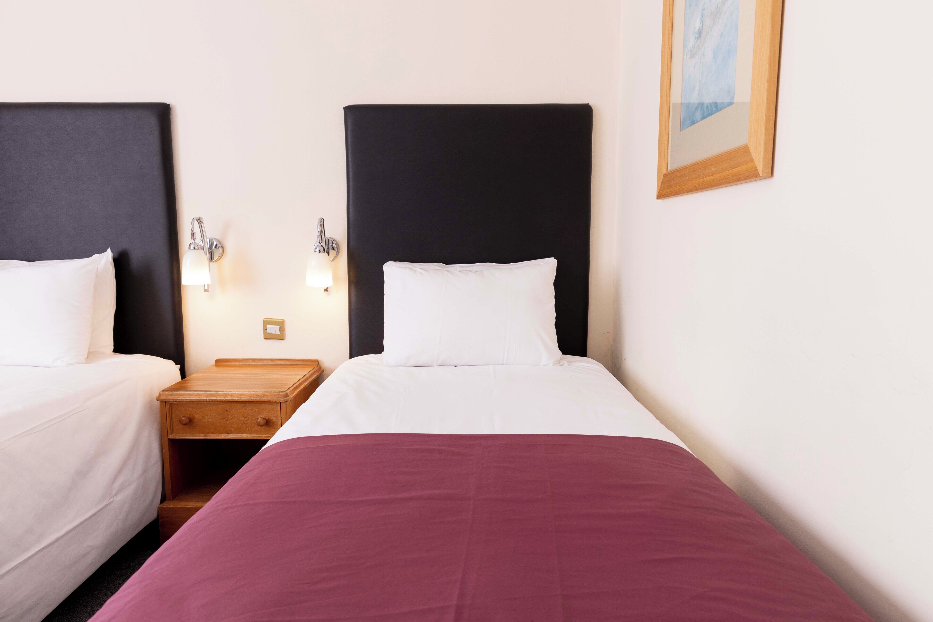 Quarto de hotel com duas camas, cada uma com cabeceira preta, roupa de cama branca e uma mesa de cabeceira de madeira com uma luminária.