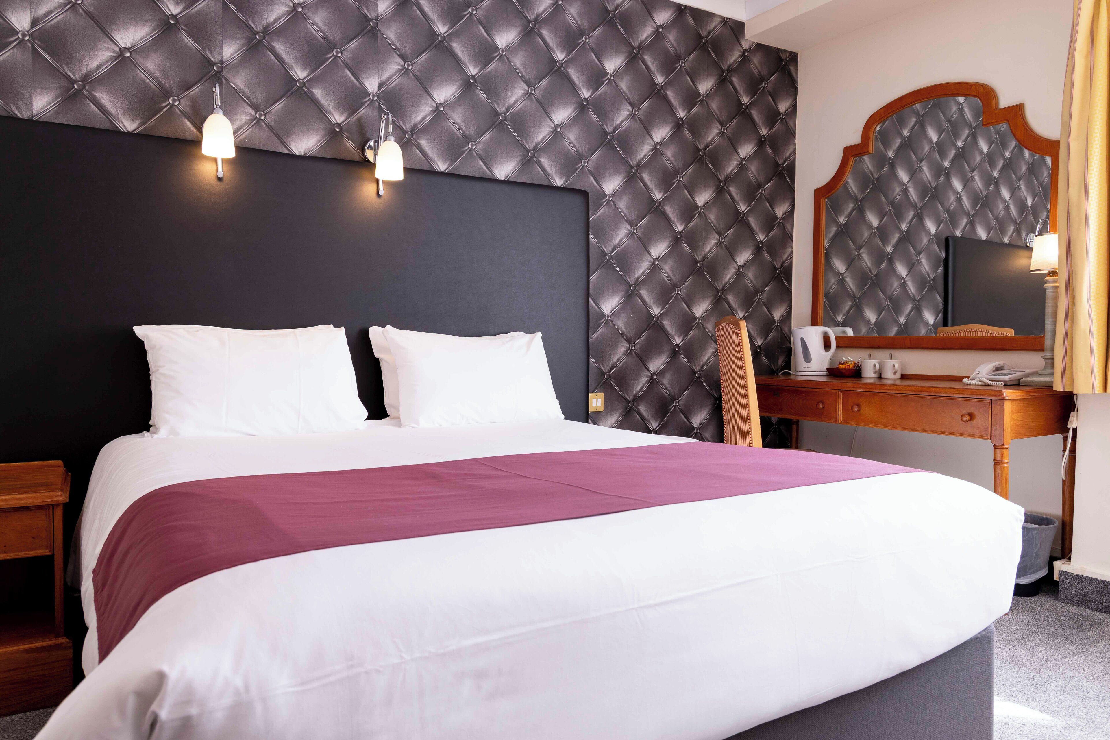 A hotel room with a large bed, a wooden desk, a chair, a television, and a wall-mounted mirror.