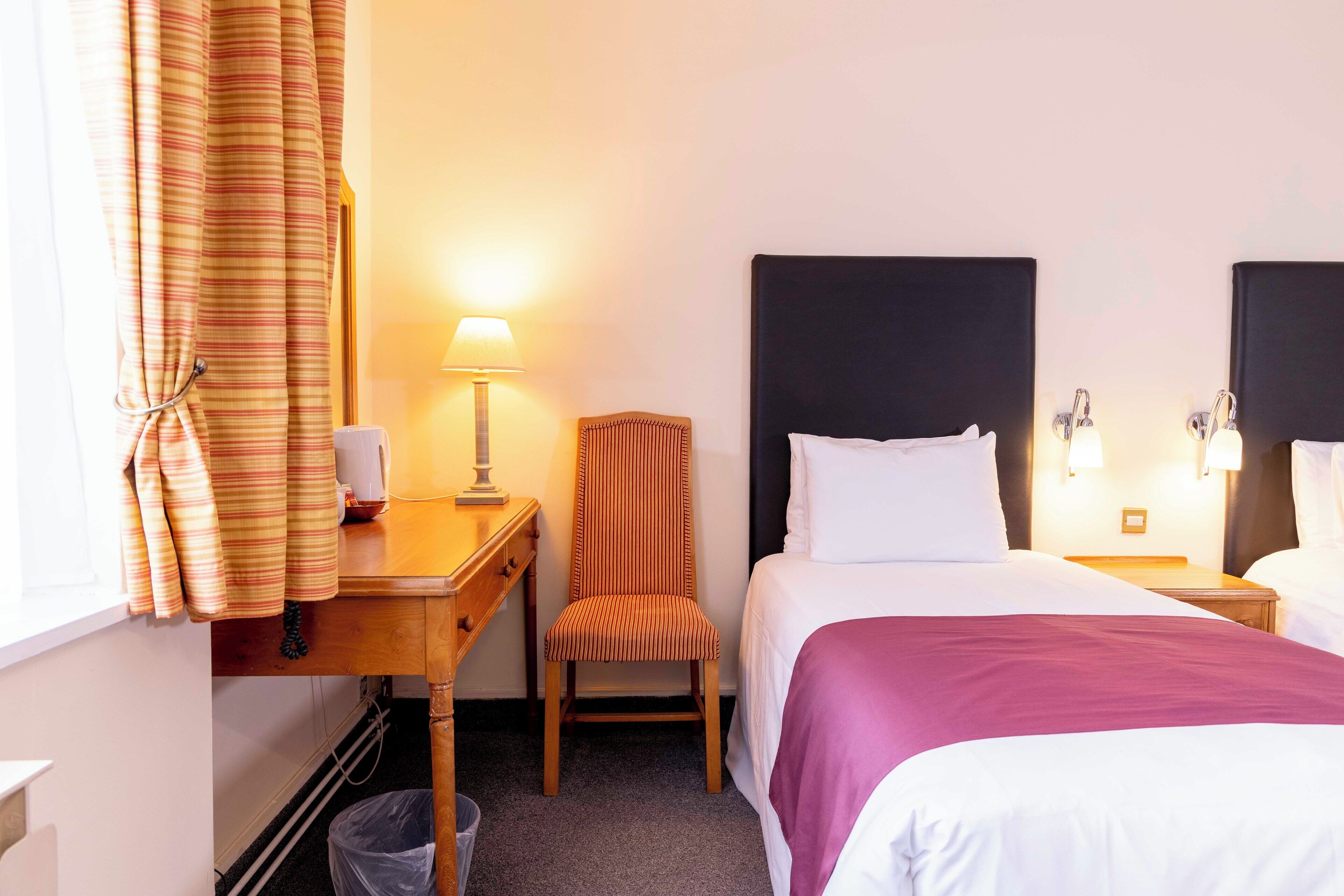 A hotel room with a bed, a chair, a desk, a lamp, and a trash bin.