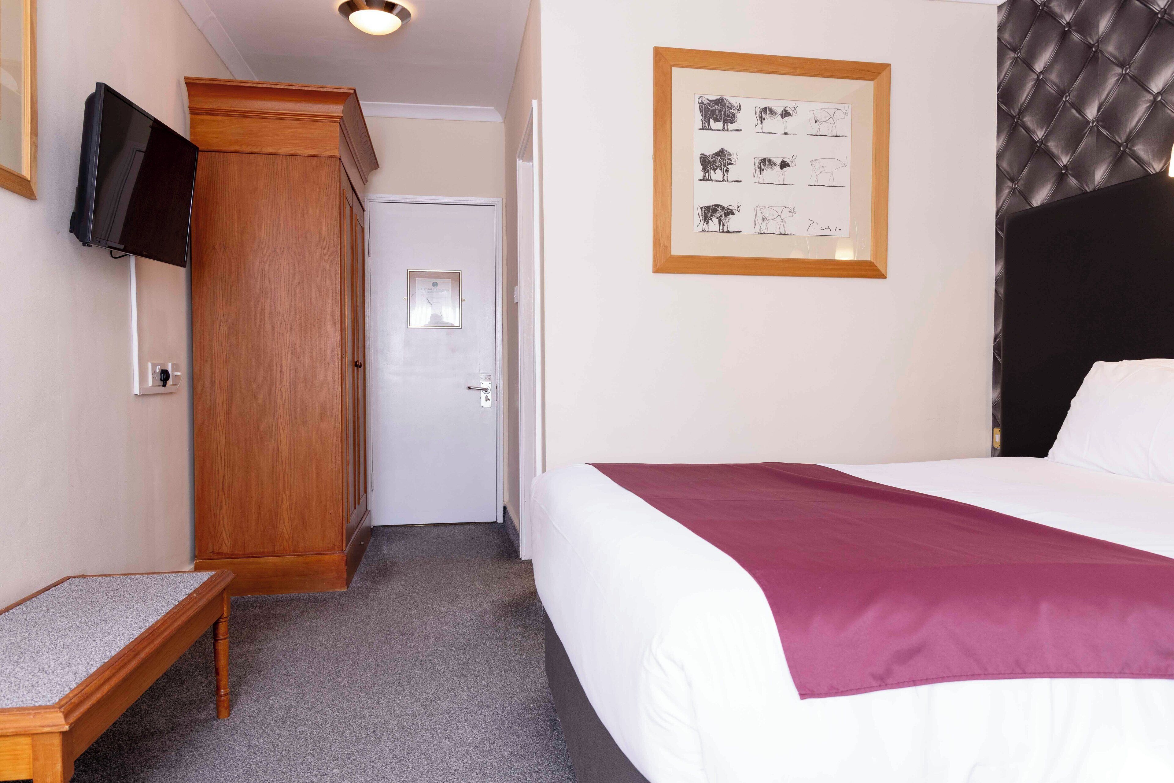 A hotel room with a bed, a wooden wardrobe, a television mounted on the wall, and a framed picture of animals on the wall.