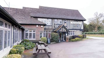 A traditional pub with a sign reading "The Roebuck." It features a stone facade, timber framing, and a gravel driveway with picnic tables.