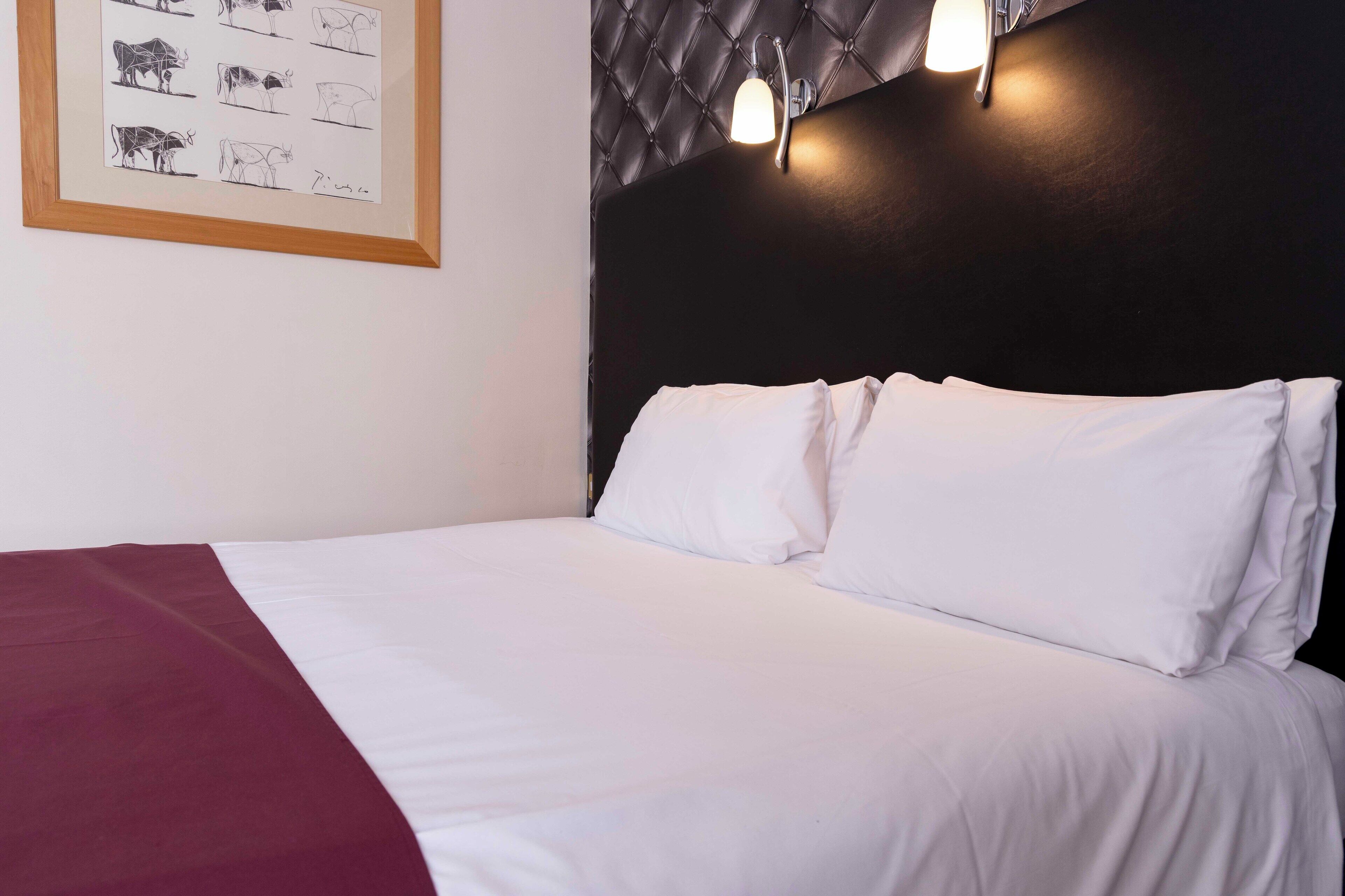 Una cama bien hecha con ropa de cama blanca y color burdeos, un cuadro enmarcado con vacas en la pared y un cabecero oscuro con luces fijadas a la pared.