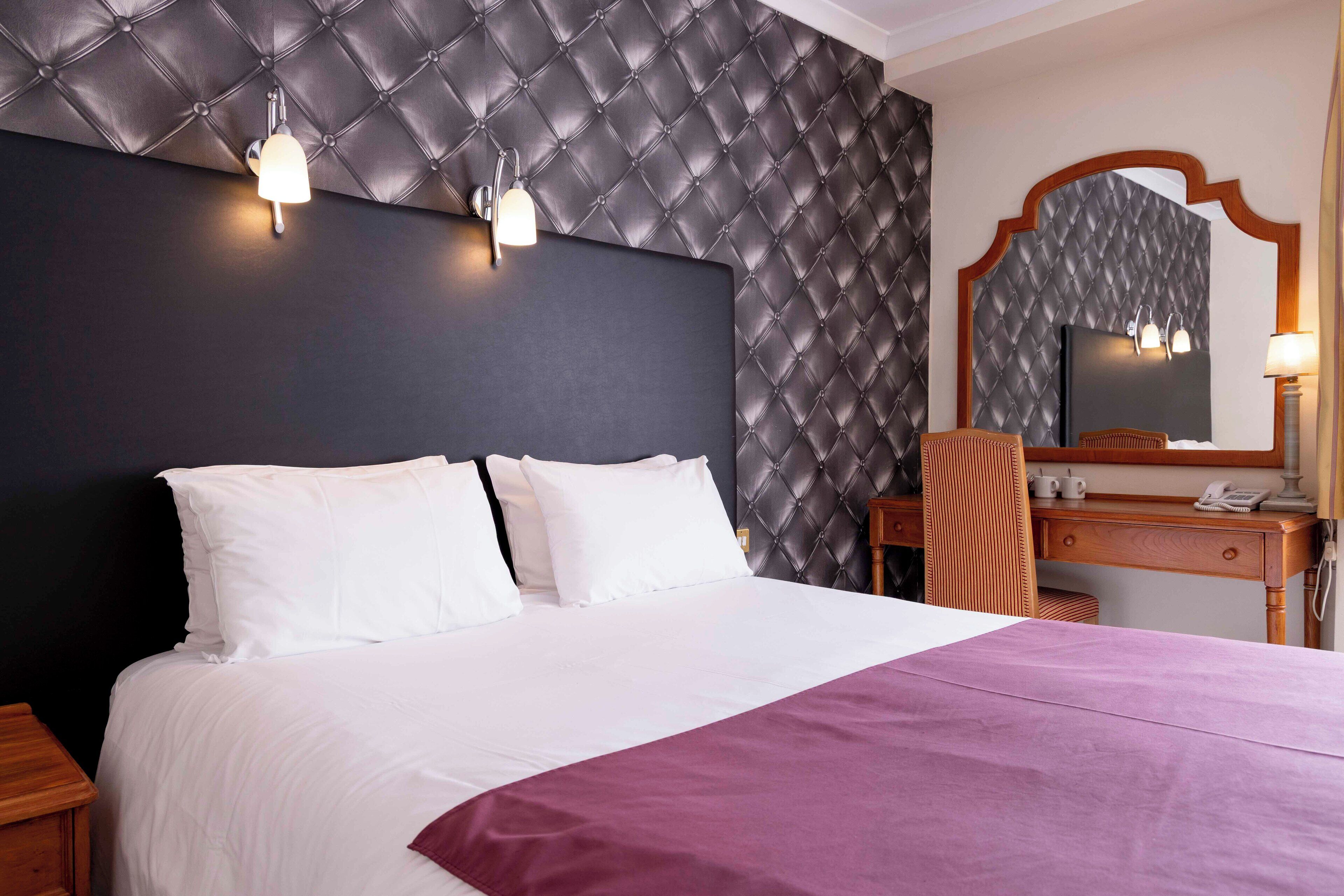 Habitación de hotel con una cama grande, una mesita de noche de madera, un escritorio con silla y un espejo.