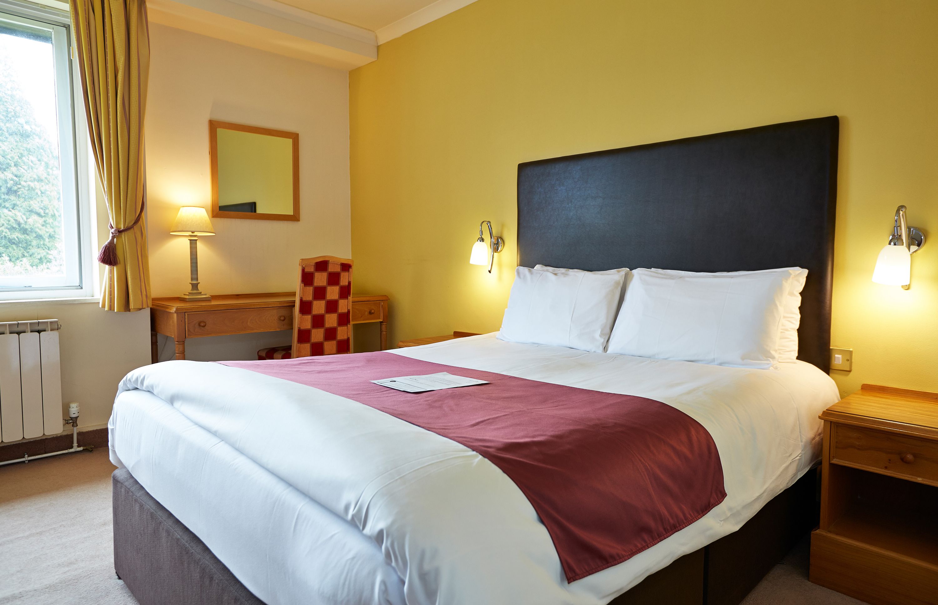 Habitació d'hotel amb un llit gran, una taula de nit, una làmpada, una cadira, un mirall i una finestra amb cortines.
