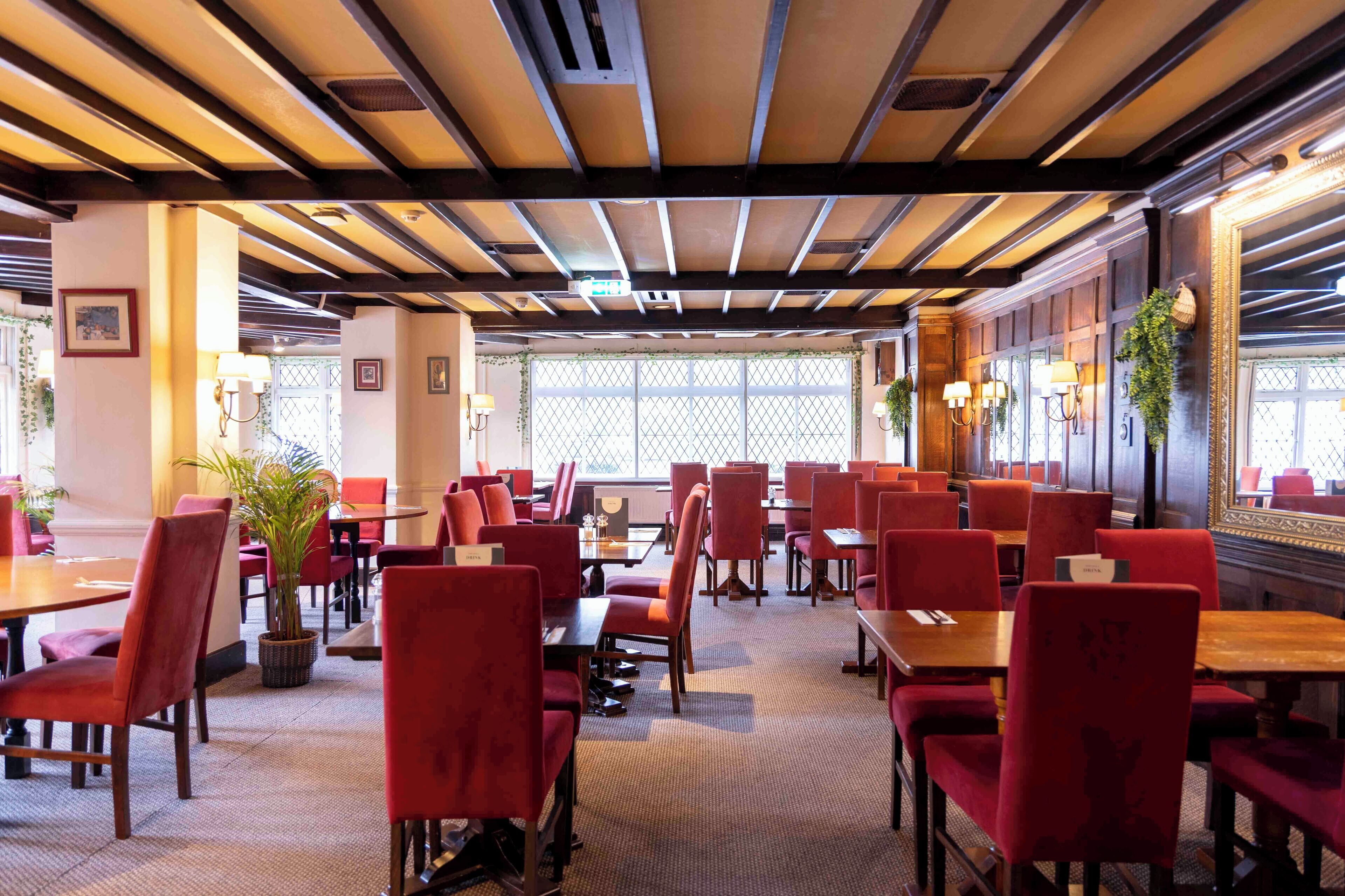 Un restaurante con sillas y mesas tapizadas de rojo, vigas de madera en el techo y grandes ventanales con vistas al exterior.