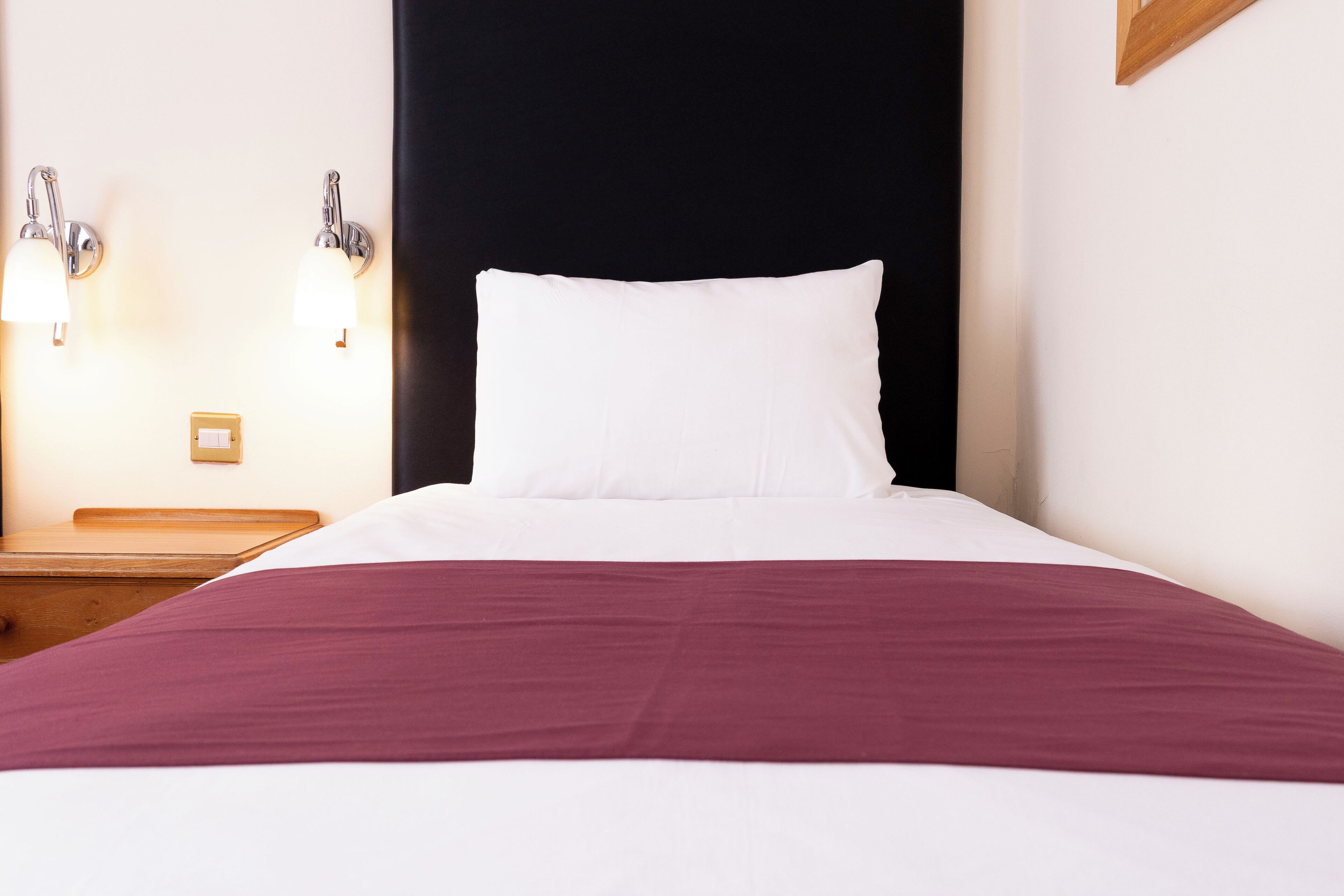 Ein ordentlich bezogenes Bett mit weißen Laken und einer bordeauxroten Tagesdecke, begleitet von zwei Wandleuchten und einem Nachttisch aus Holz.
