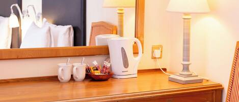 A hotel room with a wooden dresser, a mirror, two bedside lamps, a kettle, and a bowl with toiletries.