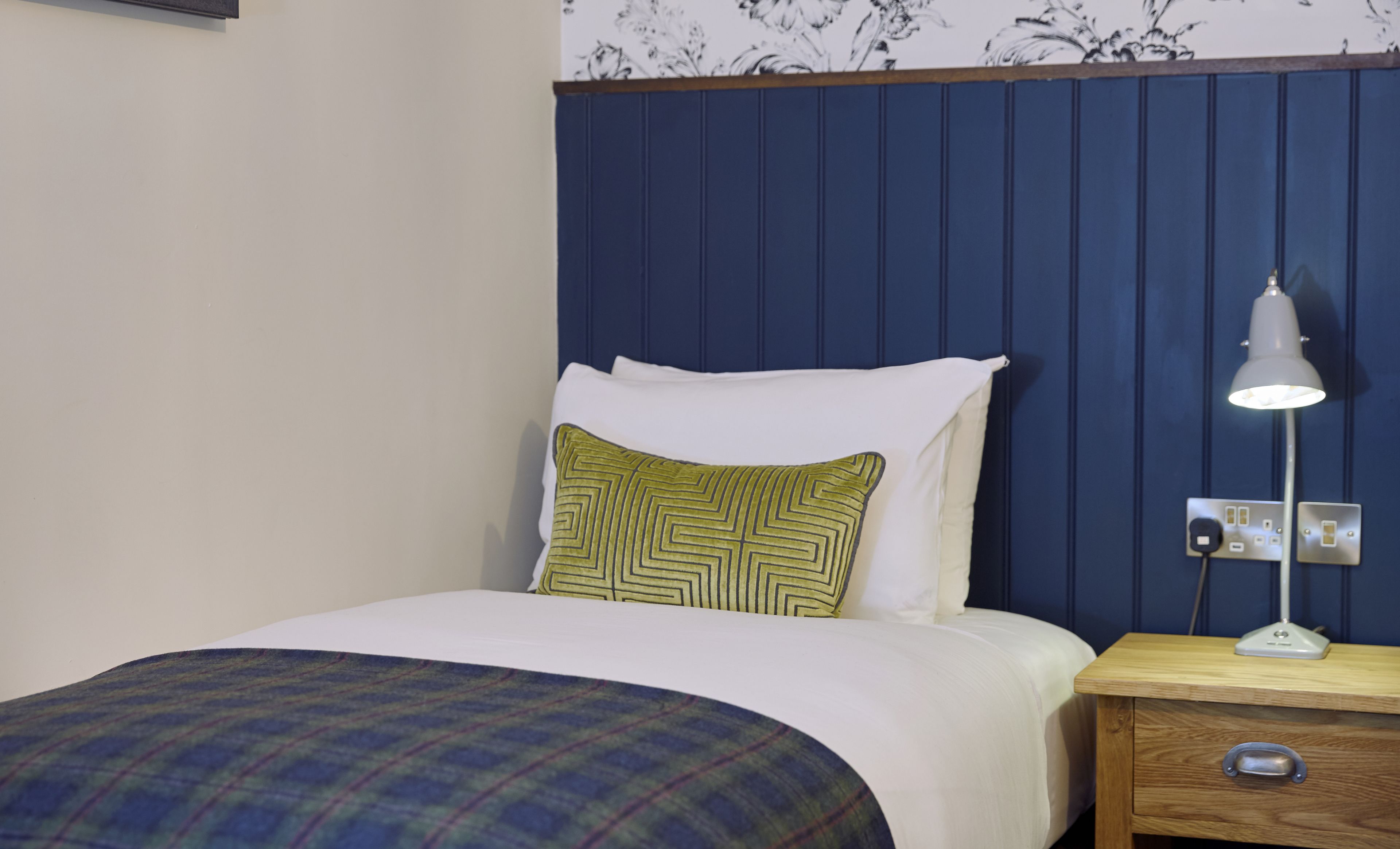 A neatly made bed with a plaid bedspread, a green and yellow patterned pillow, and a white pillow. There is a bedside table with a lamp and a power outlet.