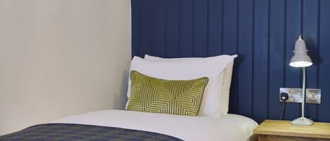 A neatly made bed with a plaid bedspread, a green and yellow patterned pillow, and a white pillow. There is a bedside table with a lamp and a power outlet.
