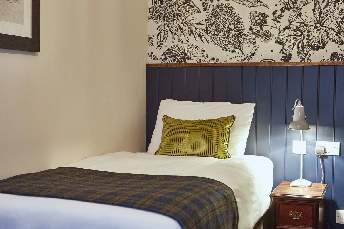 A neatly made bed with a plaid bedspread and a geometric pillow, a bedside table with a lamp, and a floral-patterned wall.
