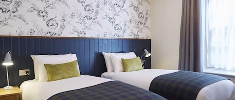 Une chambre d’hôtel avec deux lits, un papier peint à motifs floraux, une tête de lit en bois et une fenêtre avec des rideaux.