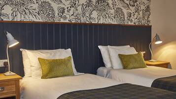 A hotel room with two beds, each with white bedding and green patterned pillows. There are bedside tables with lamps, and a floral-patterned wall above the beds.