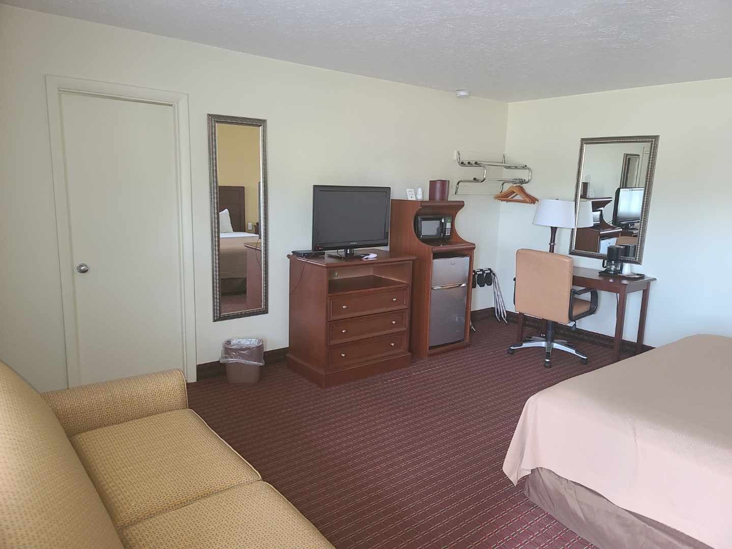 A hotel room with a bed, a sofa, a desk with a computer, a TV, a refrigerator, and a mirror.