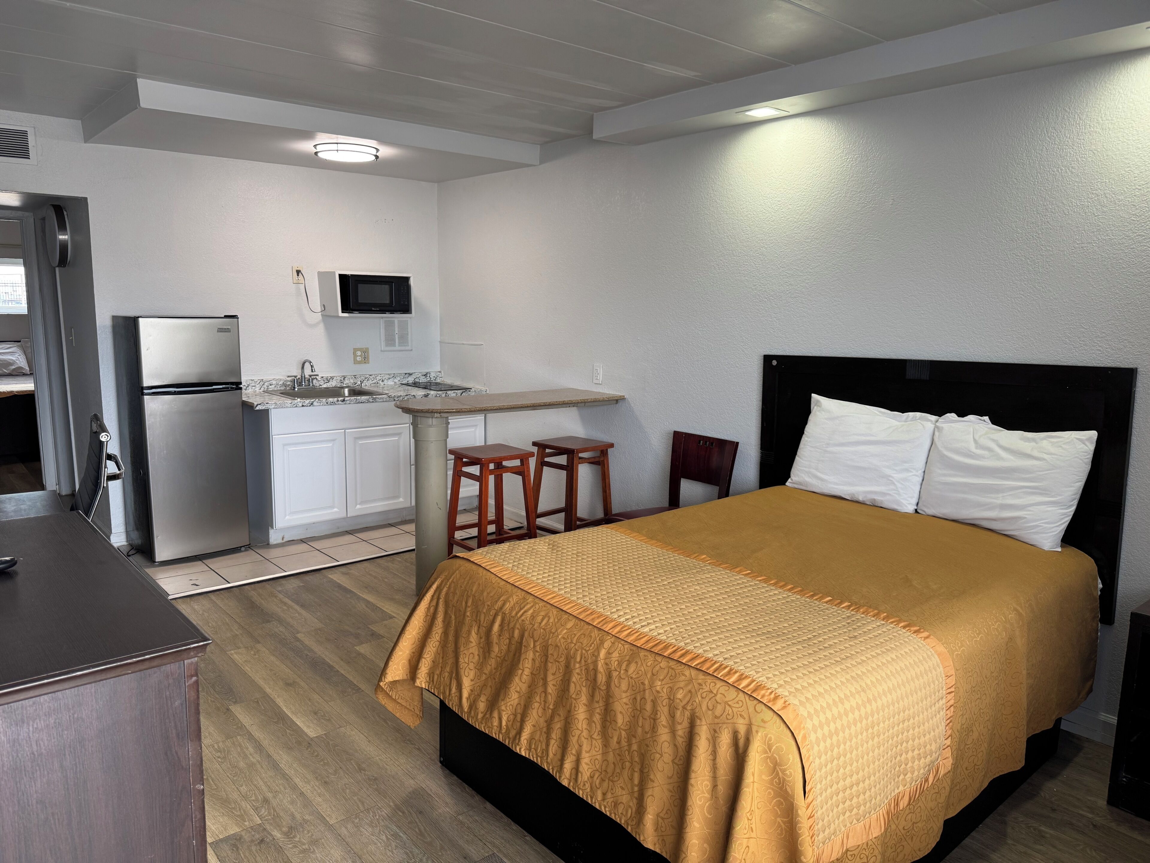 A compact hotel room with a bed, a small kitchenette, a desk, and a chair.