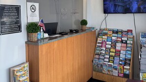 A hotel reception area with a wooden counter, a clock, a no-smoking sign, a television displaying a cityscape, and a bulletin board with various pamphlets. - Belmont Inn And Suites (Virginia Beach)