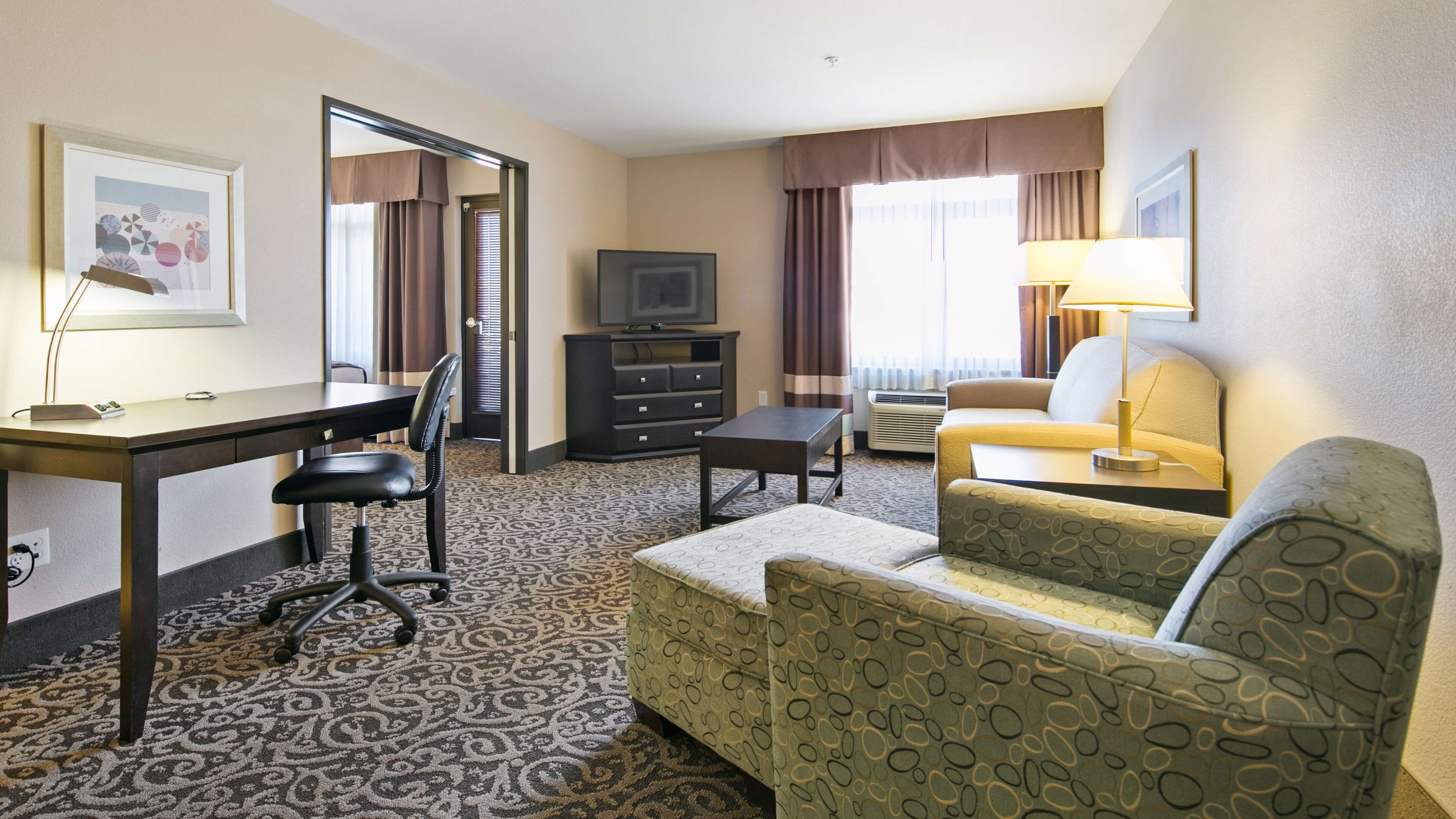 A hotel room with a desk, chair, sofa, television, and a lamp.