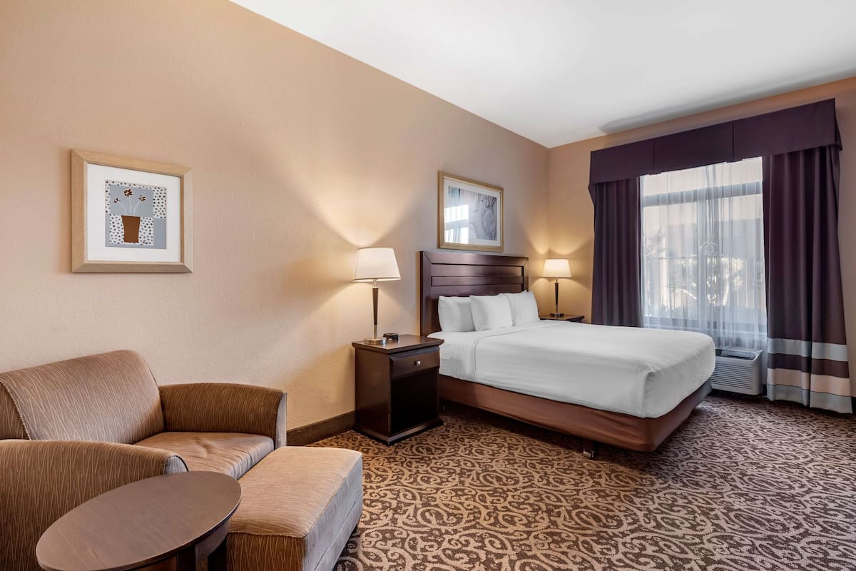 Ein Hotelzimmer mit einem Bett, einem Sessel, einem kleinen Tisch, zwei Nachttischlampen und zwei gerahmten Bildern an der Wand.