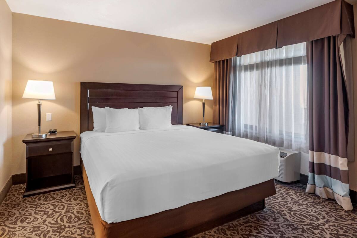 Ein Hotelzimmer mit einem großen Bett, zwei Nachttischen mit Lampen, einem Fenster mit Vorhängen und einem gemusterten Teppich.