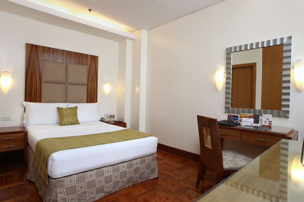 A hotel room with a bed, bedside table, desk, chair, and a large mirror.