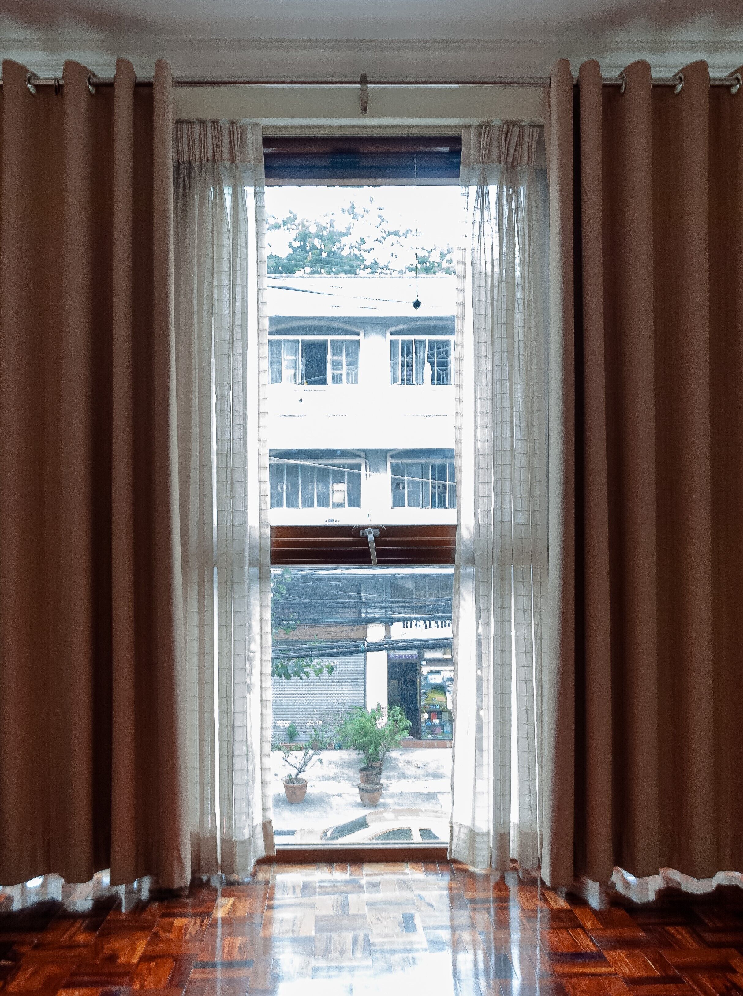 Una habitación con un ventanal grande, cortinas marrones y suelo de madera.