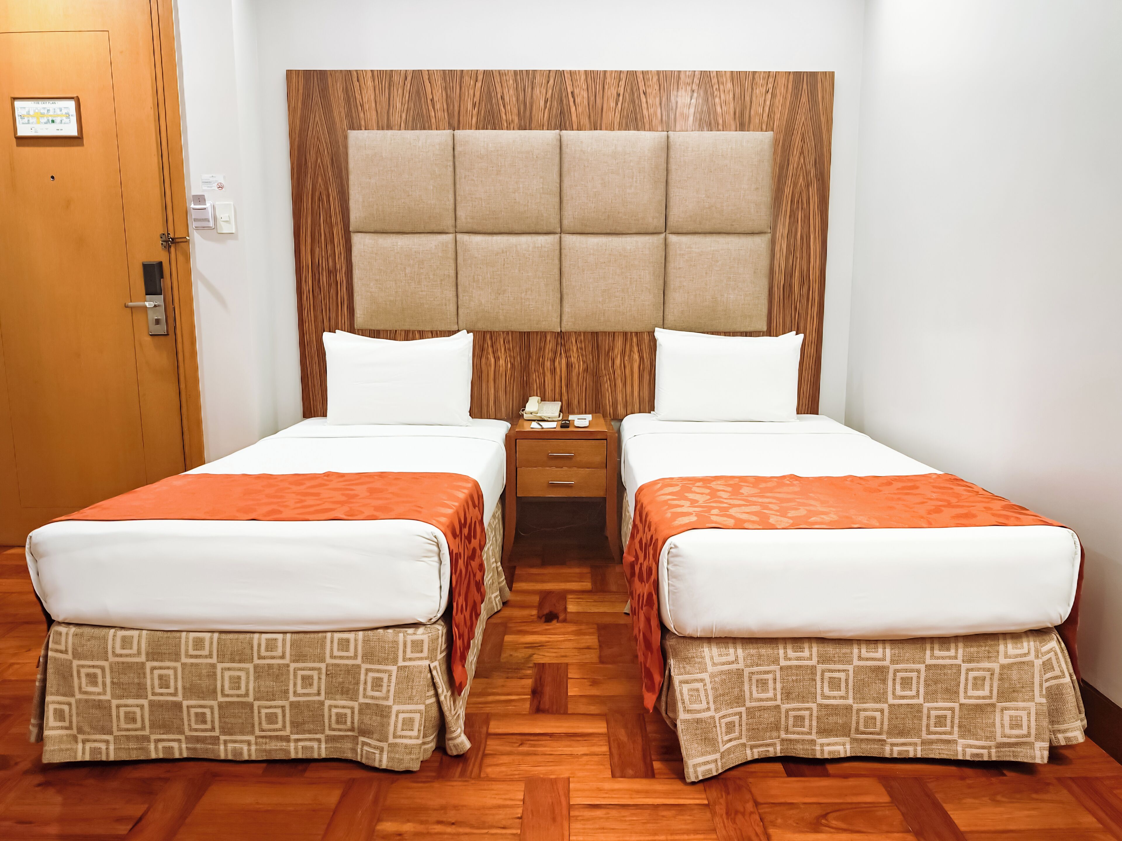 Habitación de hotel con dos camas, cabecera de madera, suelo de madera, una mesita de noche y una puerta con un cartel.