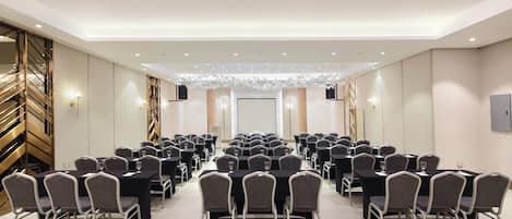A conference room with rows of tables and chairs, a stage at the front, and a decorative ceiling.
