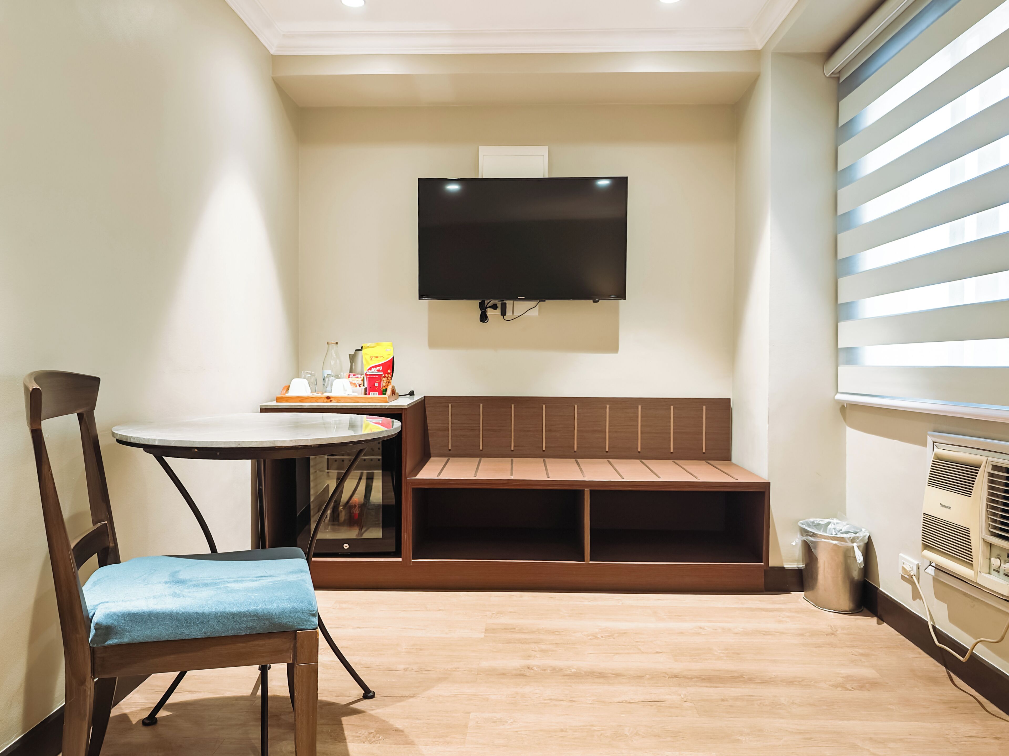 Une pièce comprenant un banc en bois, une table avec une chaise, un téléviseur fixé au mur et une fenêtre avec des stores.