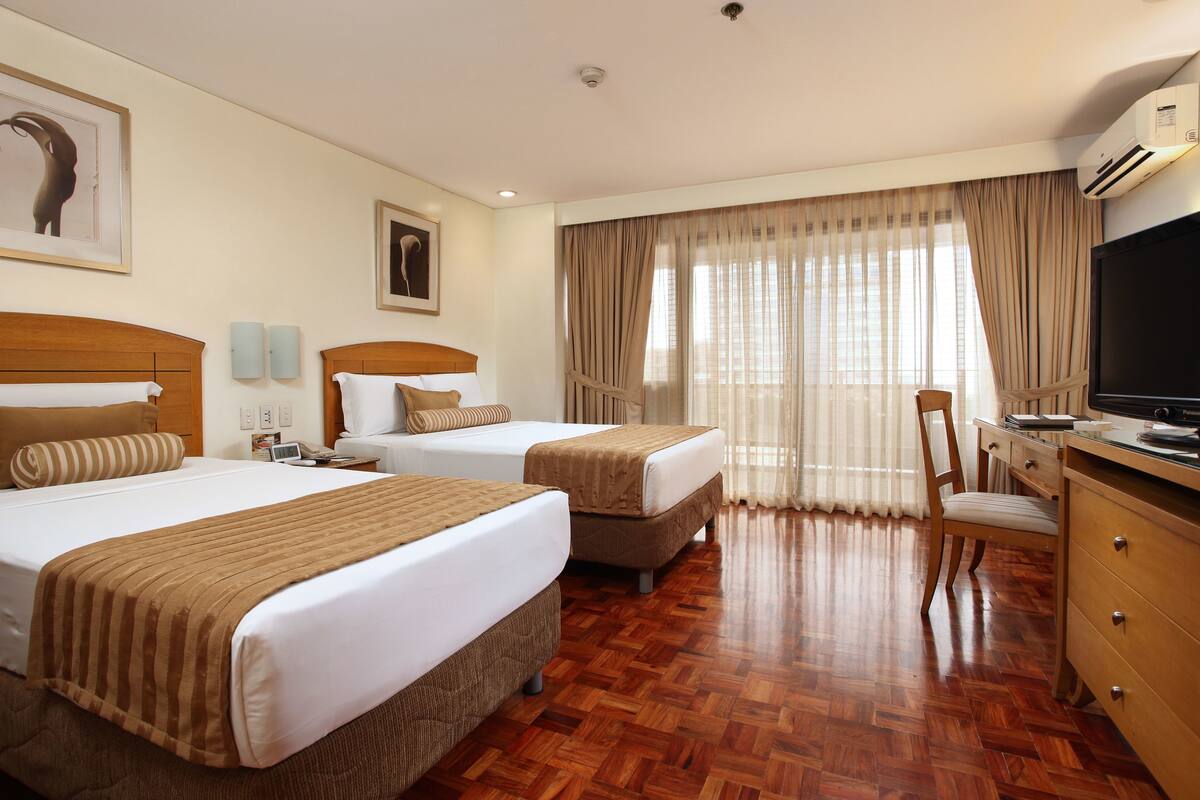 A hotel room with two beds, a desk with a TV, a chair, and a large window with curtains.