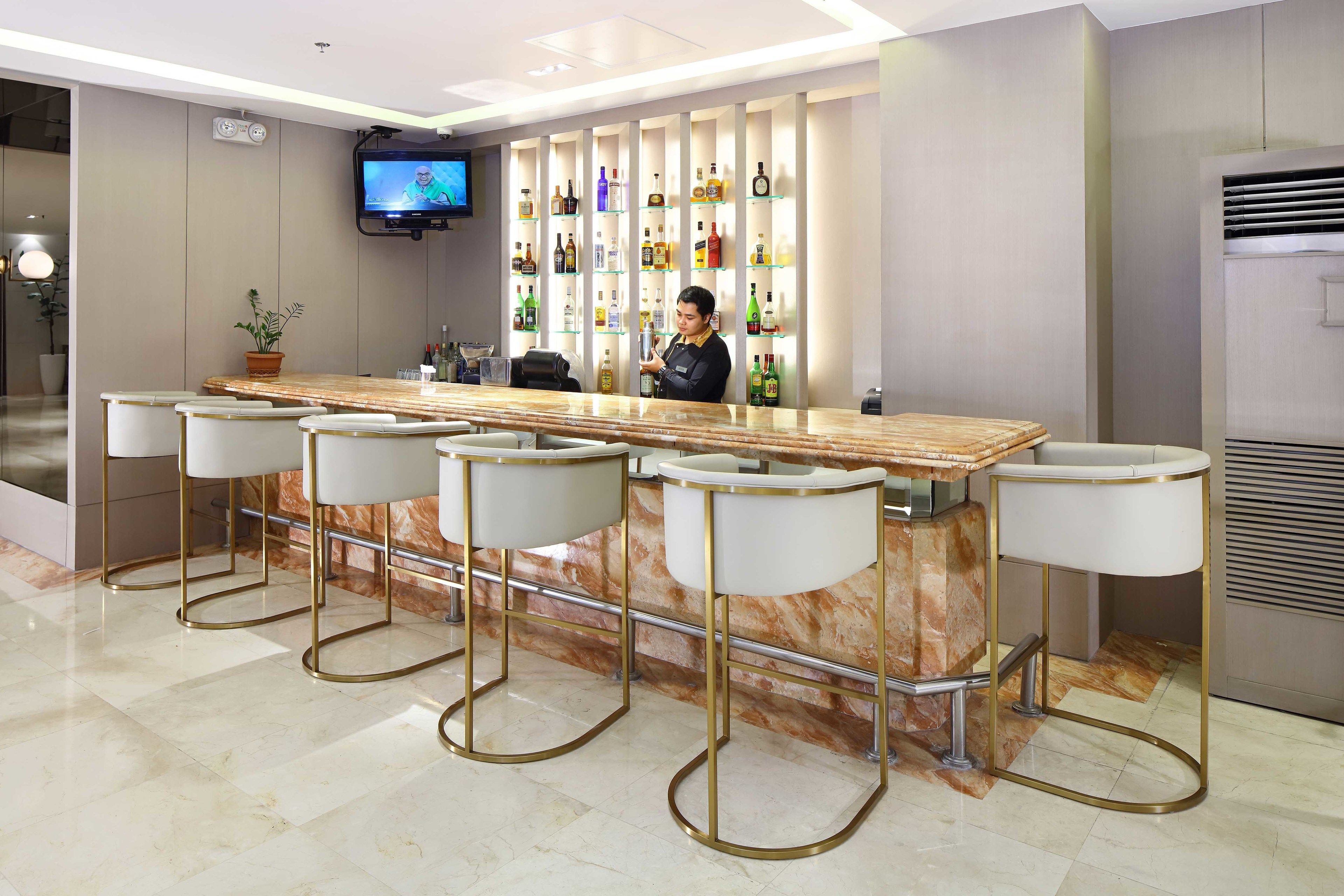 A bar area with a marble countertop and four bar stools, a television mounted on the wall, and a variety of bottles on shelves behind the bar.