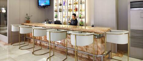A bar area with a marble countertop and four bar stools, a television mounted on the wall, and a variety of bottles on shelves behind the bar.