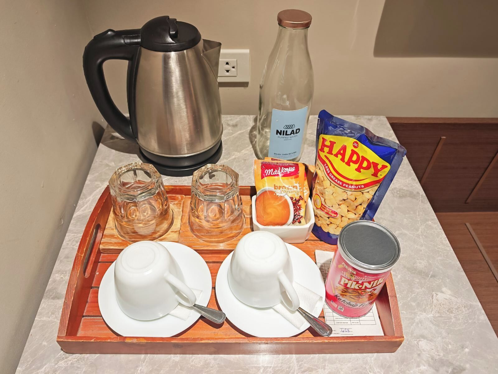 A tray with a coffee pot, a bottle, a bag of peanuts, a bowl of soup, two cups, and a can of ketchup.