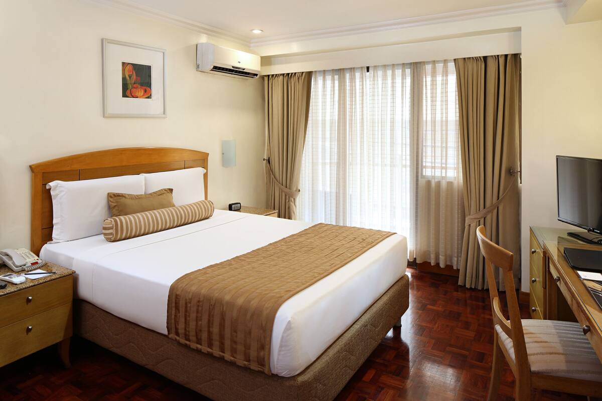 A hotel room with a large bed, a wooden headboard, a desk with a chair, a television, and a window with curtains.