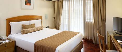 Habitación de hotel con una cama grande, un cabecero de madera, un escritorio con una silla, un televisor y una ventana con cortinas.