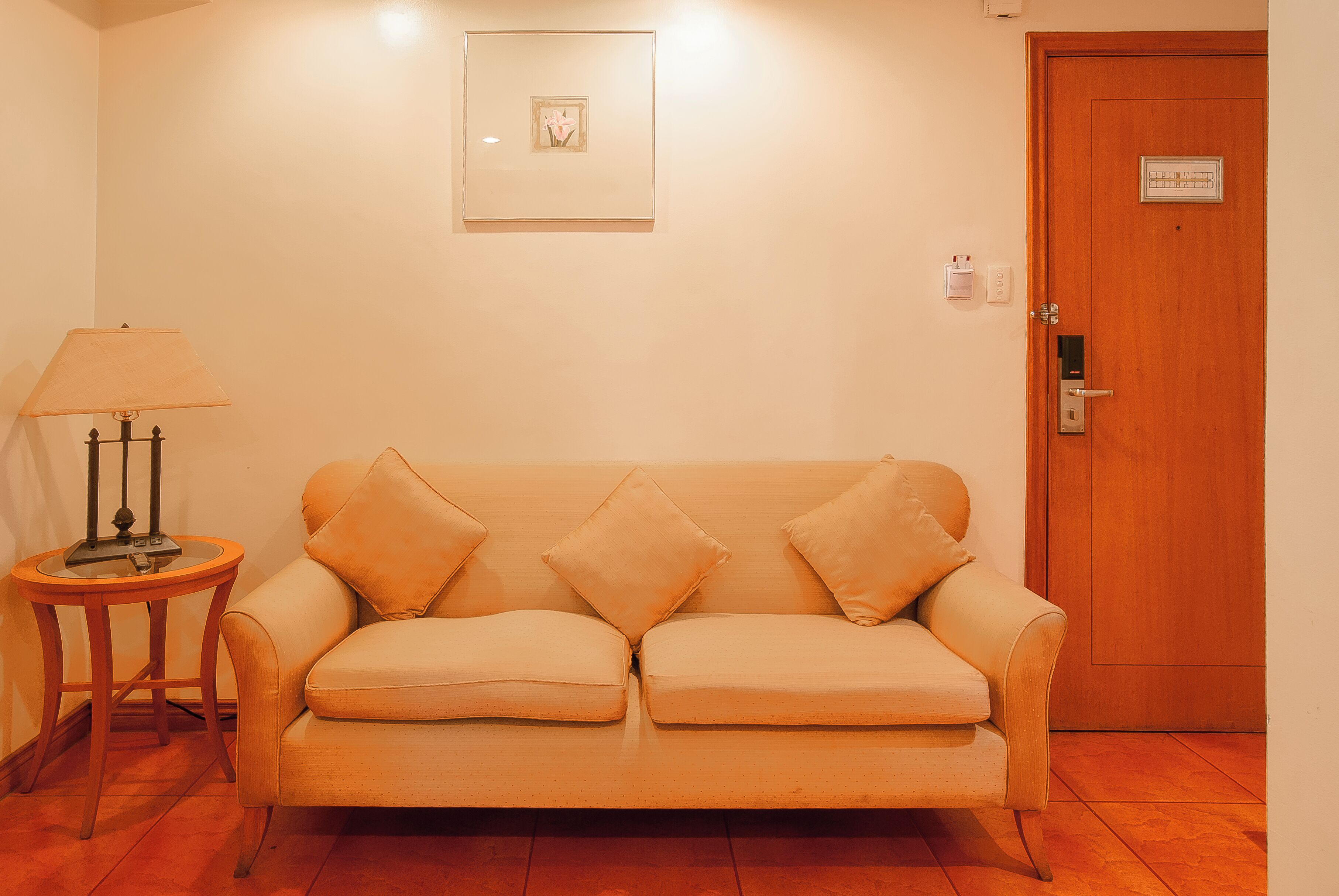 A room with a beige sofa, a small table with a lamp, a framed picture on the wall, and a wooden door with a lock.