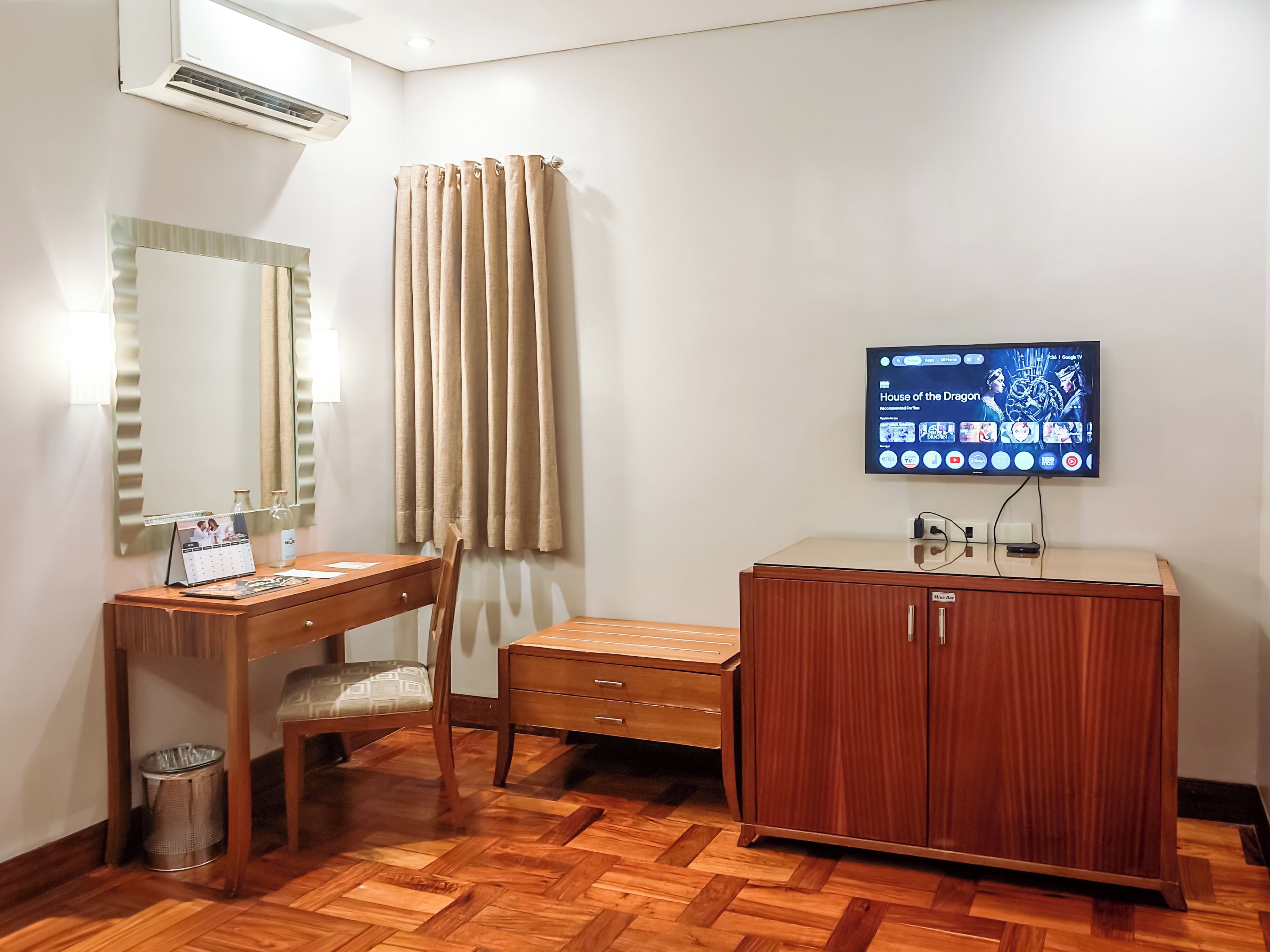 A hotel room with a wooden desk, chair, and cabinet, a mirror, a television mounted on the wall, and a window with curtains.