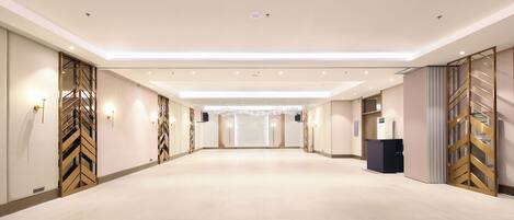 A spacious hotel corridor with a modern design, featuring a long, open layout with a central light fixture and a podium at one end.