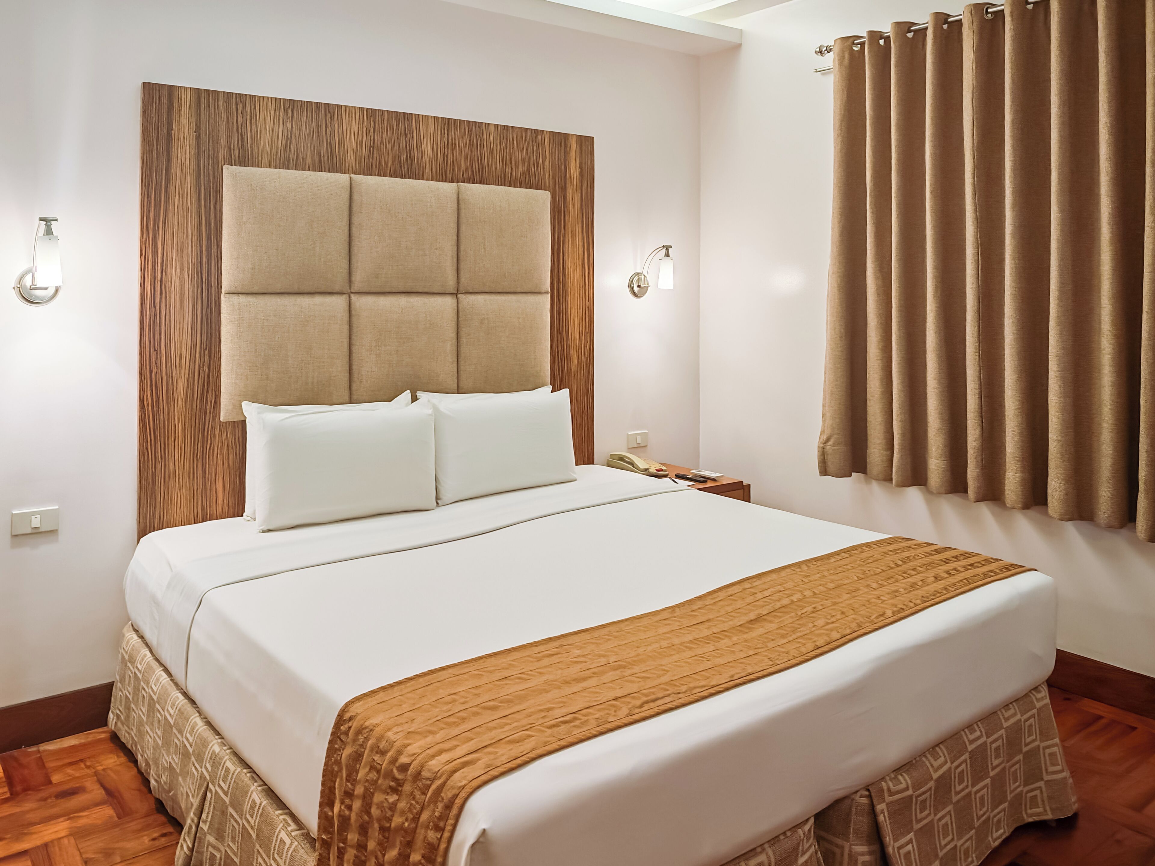 A hotel room with a large bed, a wooden headboard, a bedside table with a lamp, and a window with curtains.