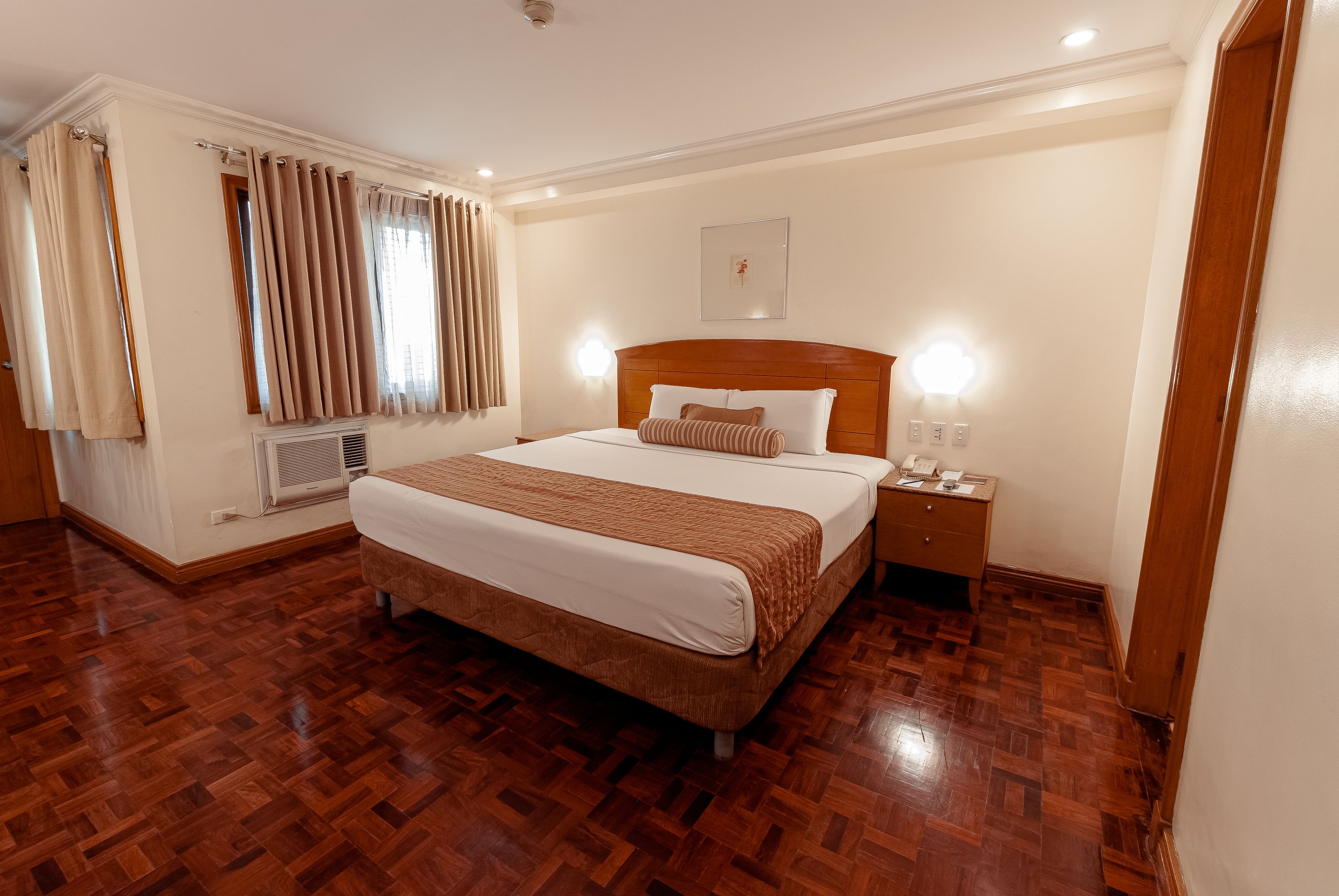 Una habitación de hotel con suelo de madera, una cama grande con cabecero, una mesita de noche y un buró.