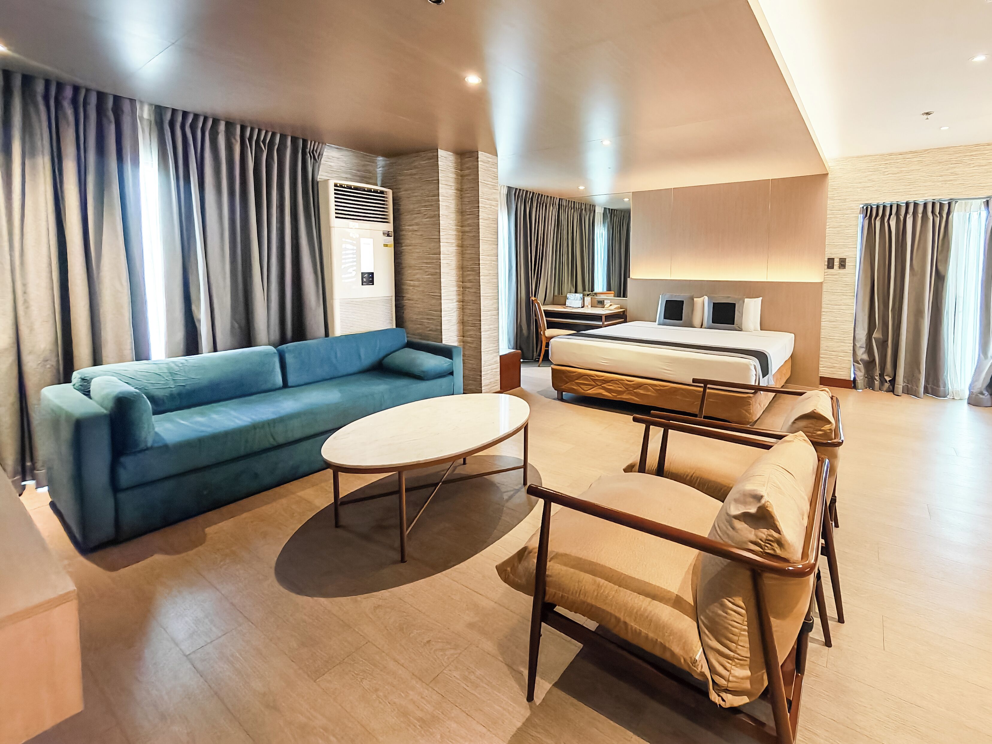 A modern hotel room with a blue sofa, a round table, a bed with a wooden frame, and a desk with a chair.