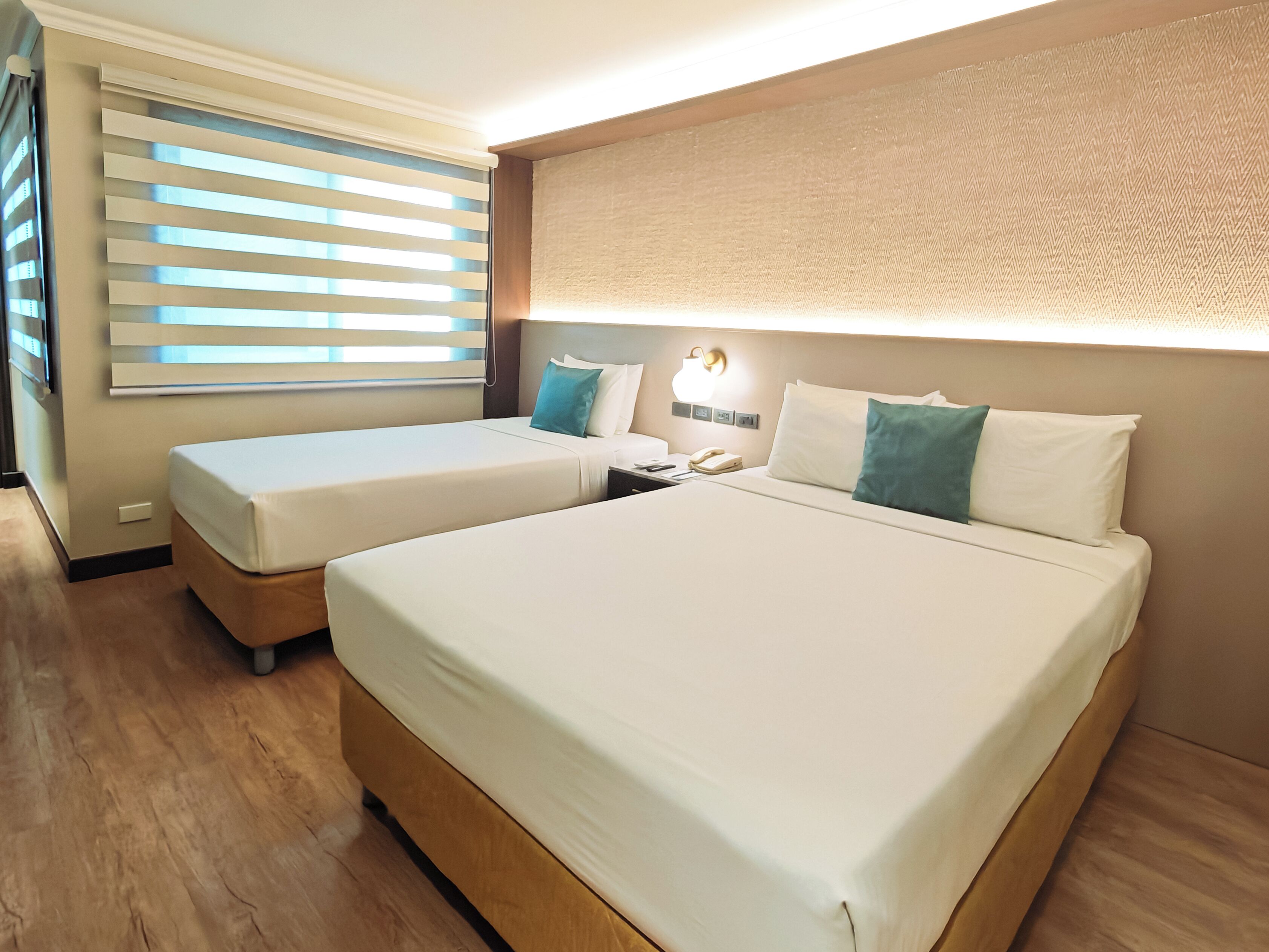 A hotel room with two beds, a bedside table with a lamp, and a window with blinds.