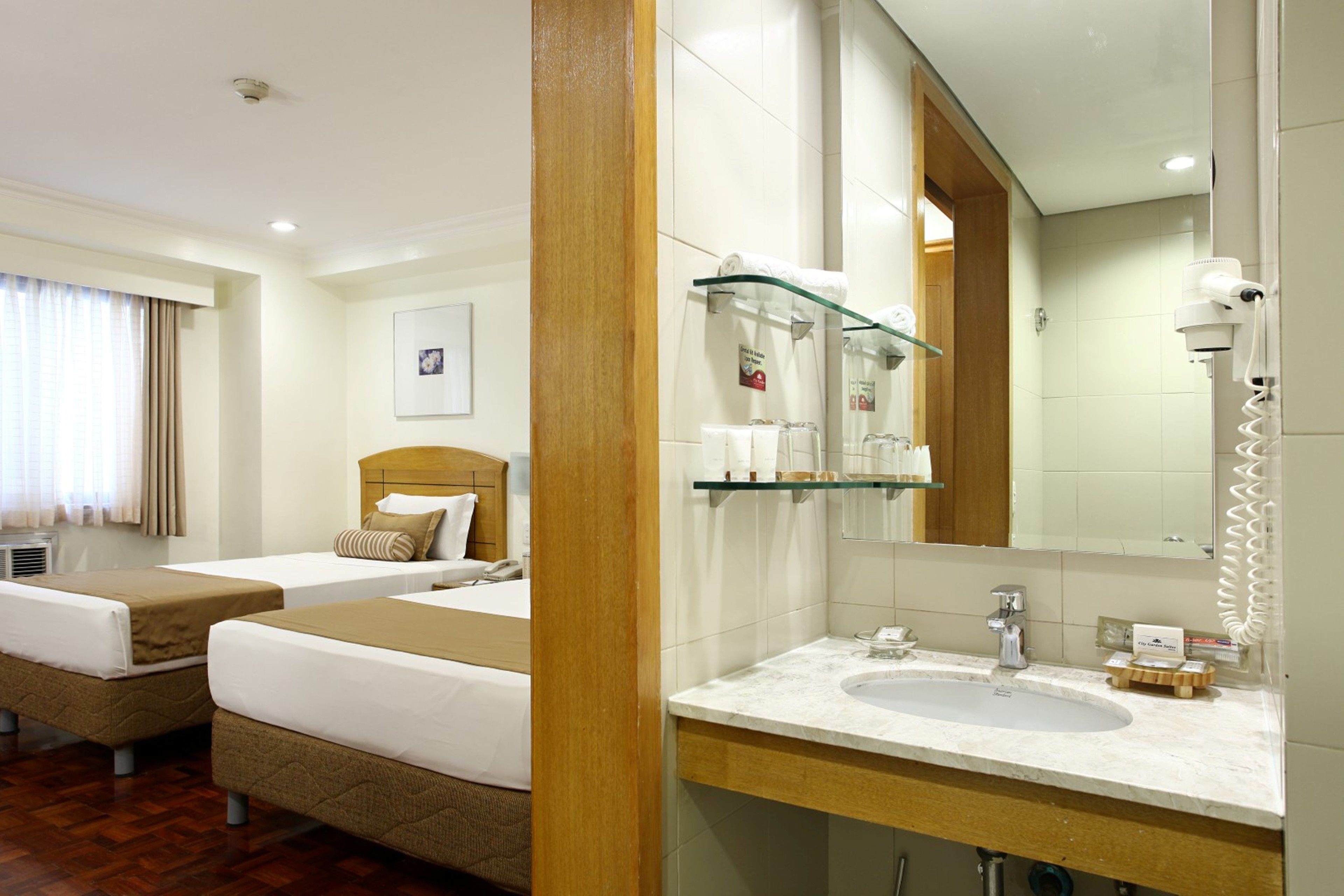 A hotel room with two beds, a large mirror, a bathroom with a sink and toiletries, and a shower area.