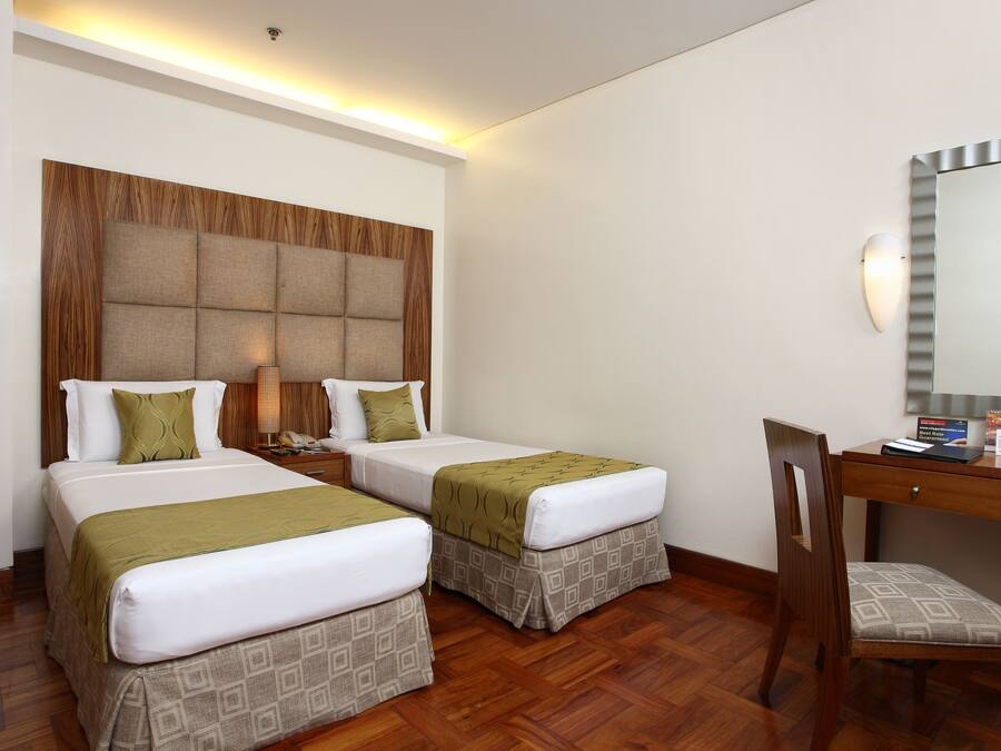 Quarto de hotel com duas camas, uma cabeceira de madeira, uma escrivaninha com cadeira e um espelho.