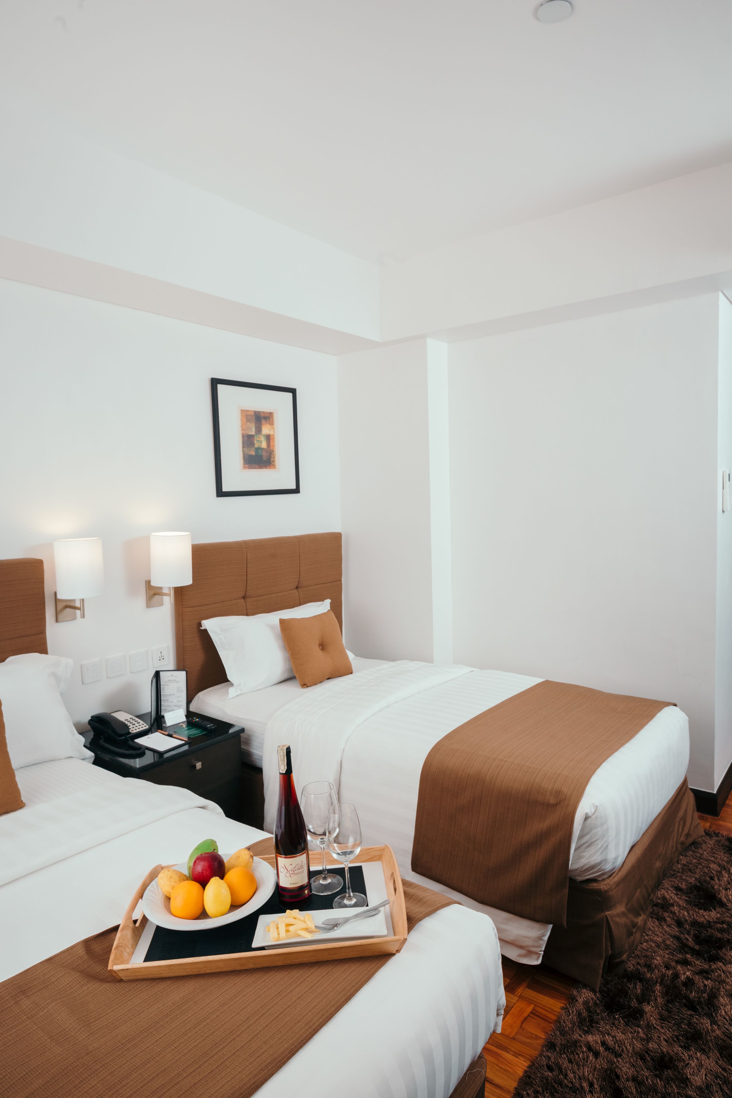 A hotel room with two beds, a bedside table with a phone and a tray with fruit and drinks, and a framed picture on the wall.