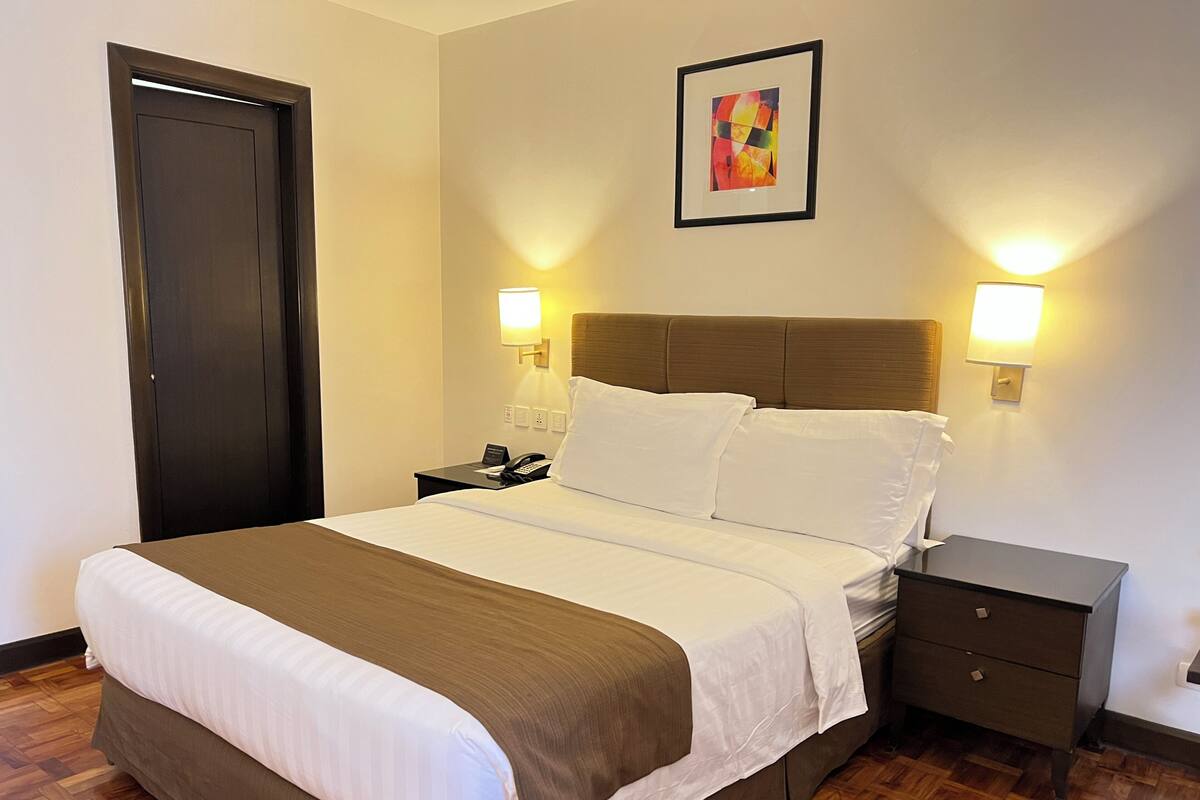 Habitación de hotel con cama, mesitas de noche, una puerta y un cuadro en la pared.