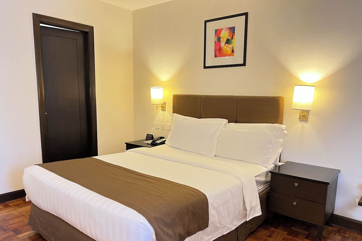 Habitació d'hotel amb llit, taules de nit, una porta i un quadre a la paret.