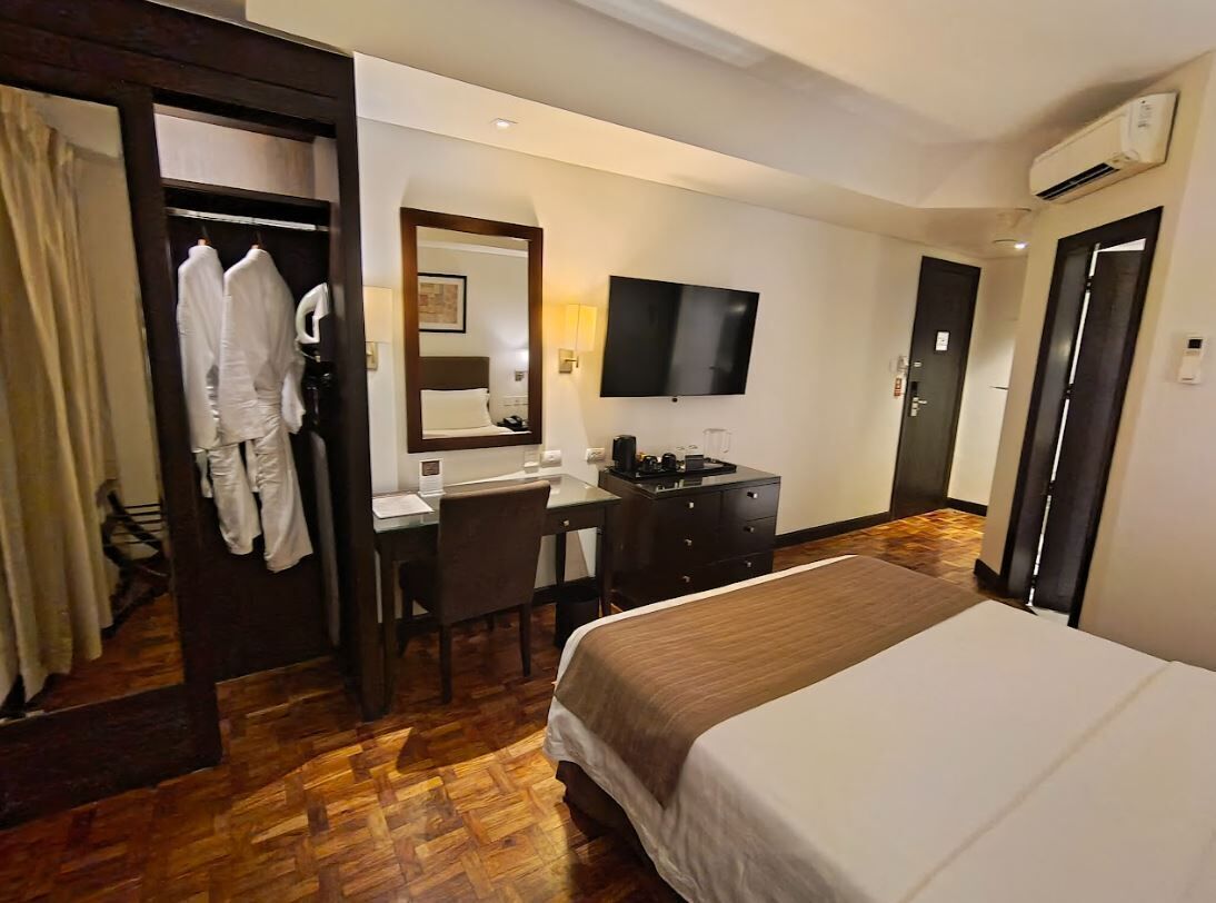 Una habitación de hotel con una cama, un escritorio, una silla, un espejo, un televisor y un armario.